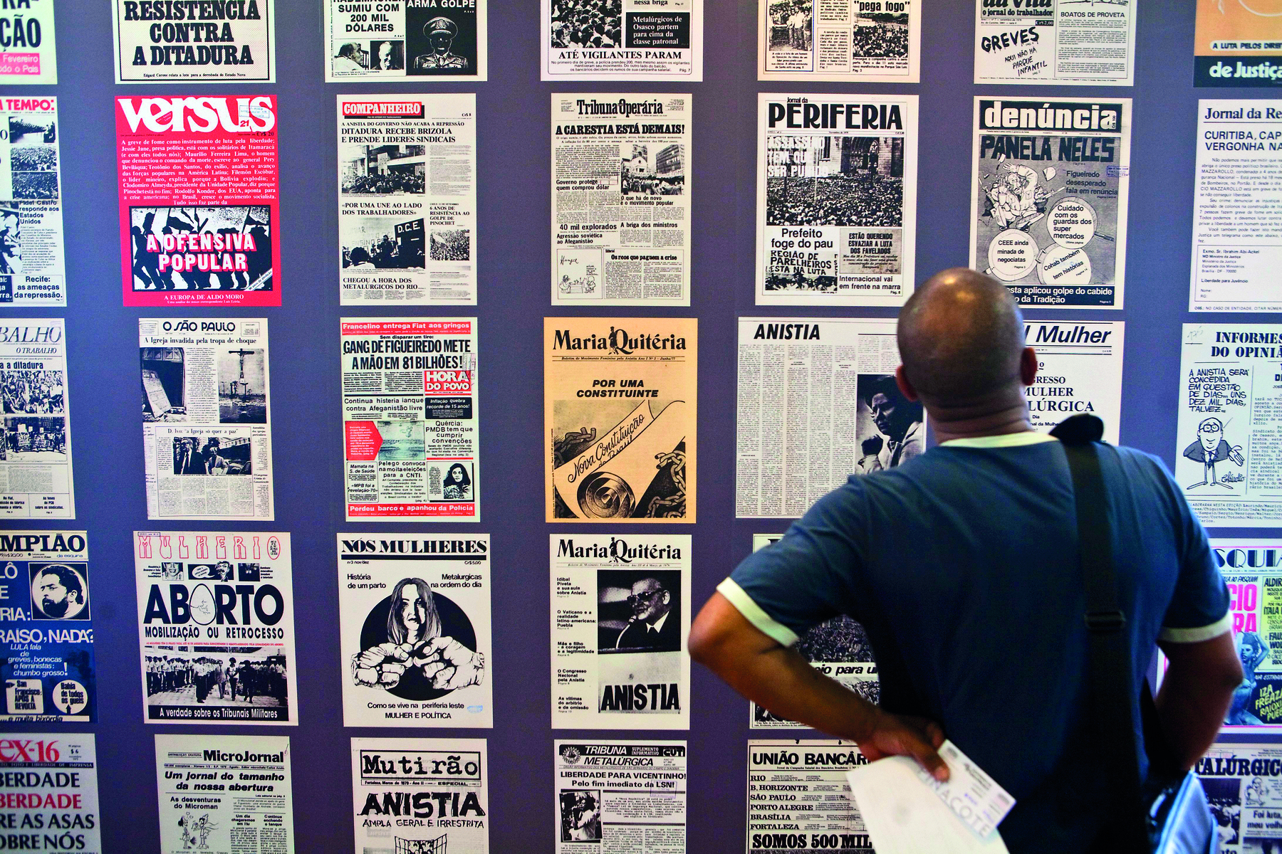 Fotografia. Homem de costas observando um mural de jornais e revistas antigas. Ele é calvo e veste camisa azul, com uma bolsa pendurada no ombro.
