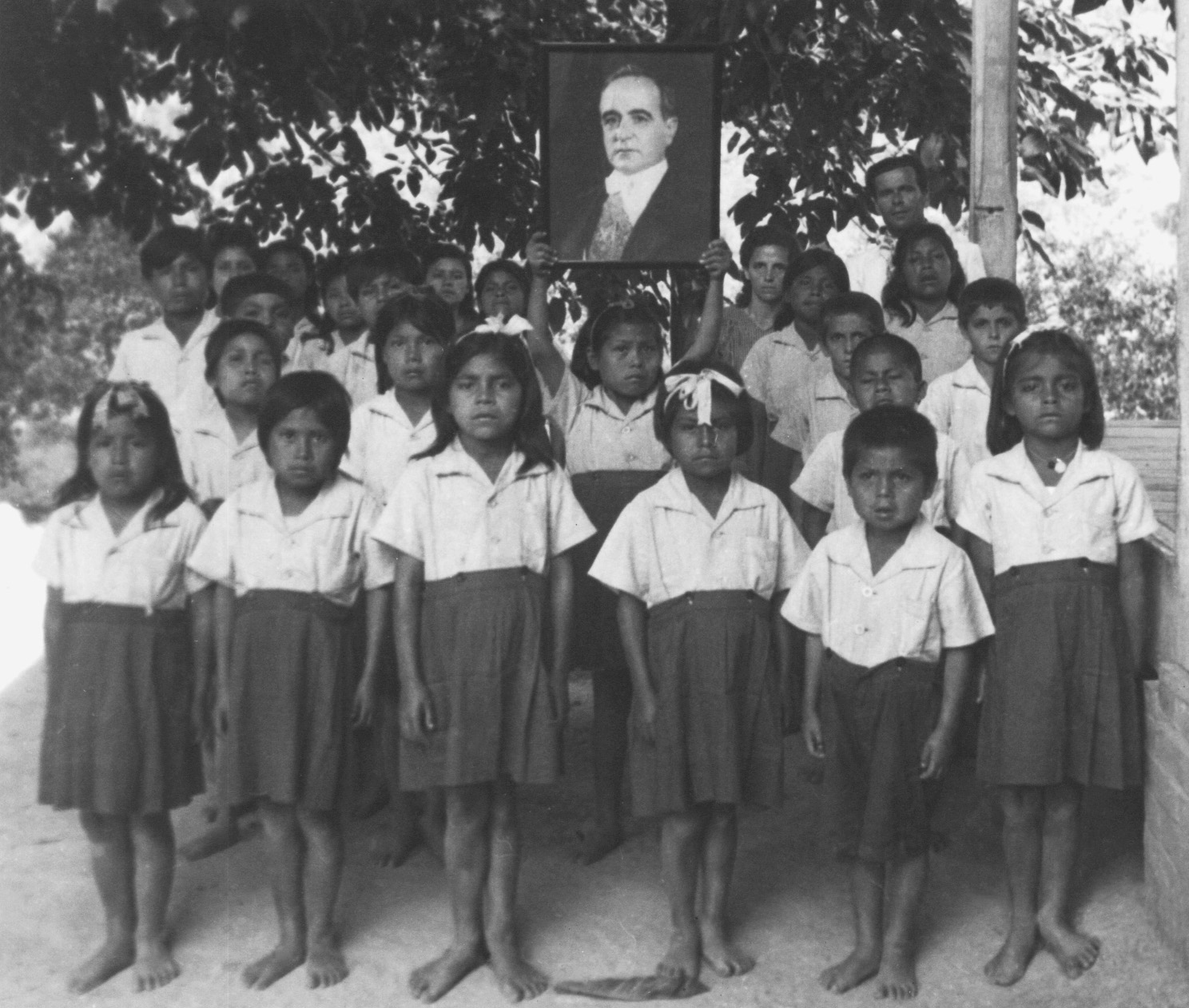 Fotografia em preto e branco. Crianças indígenas posando para foto. Estão de uniforme. Camisa clara e saia ou bermuda escura e larga. A criança no centro do grupo ergue um retrato de Getúlio Vargas.