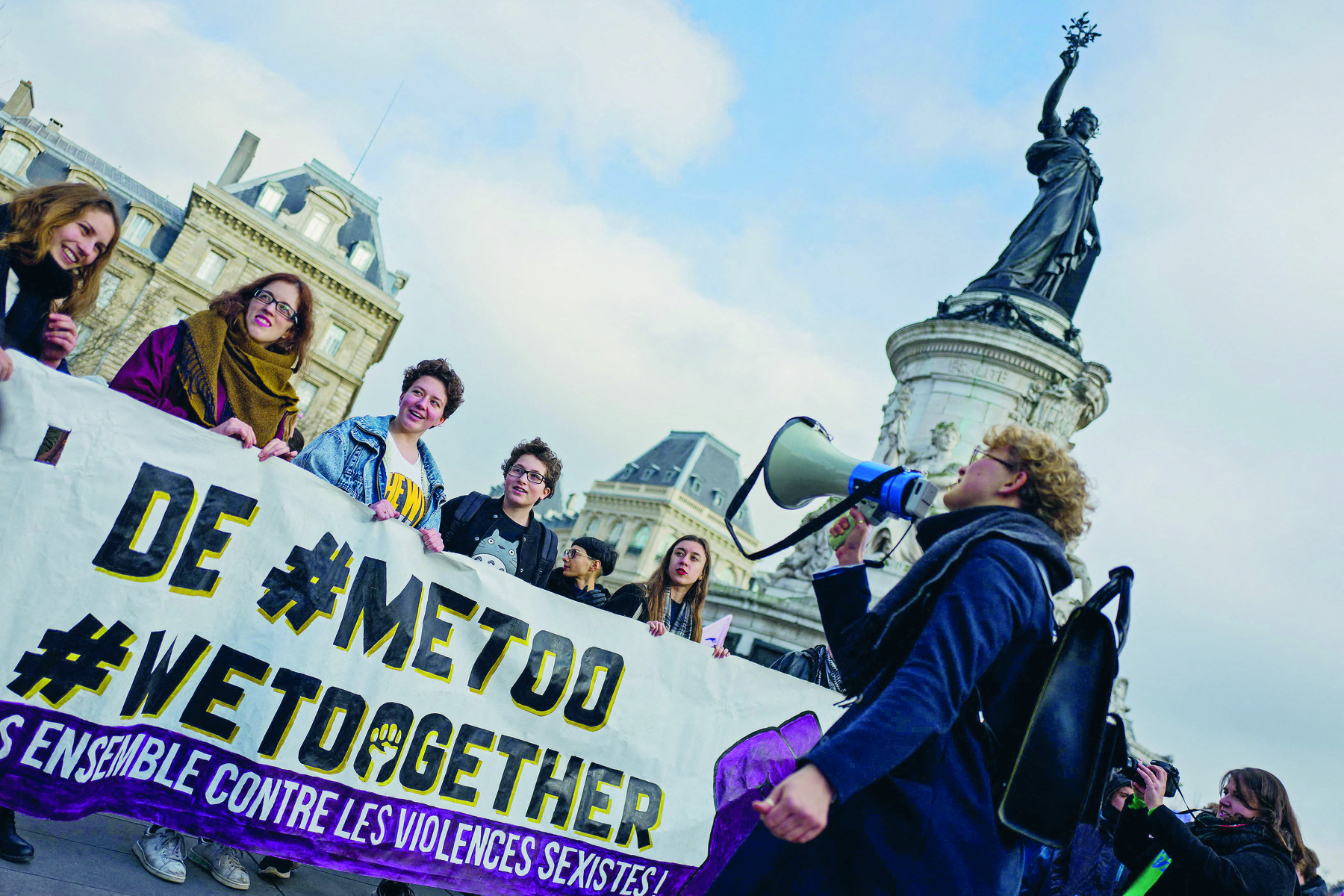 Fotografia. Mulheres em uma manifestação. Carregam juntas uma faixa com os dizeres: DE #METOO #WETOGETHER. Na frente da faixa uma pessoa falando no megafone.