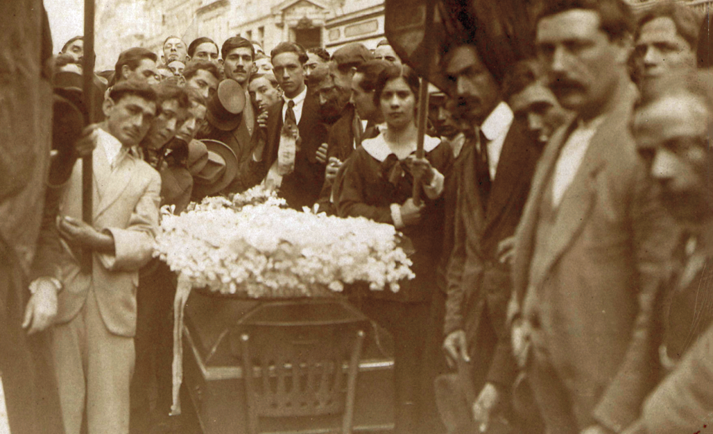 Fotografia. Homens e mulheres vestidos socialmente, aglomerados ao redor de um caixão. Sobre ele, flores claras.