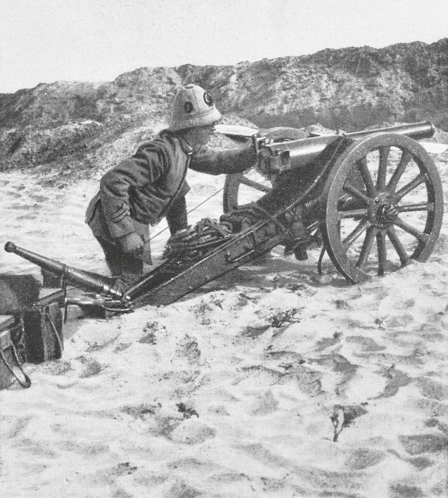 Fotografia em preto e branco. Soldado ajoelhado na praia, manuseando um pequeno canhão. Ele está apoiado em uma estrutura com duas rodas grandes.