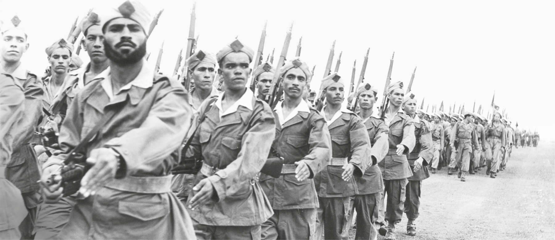 Fotografia em preto e branco. Soldados perfilados em uma longa fila. Estão com os braços em movimento e carregam baionetas sobre os ombros.