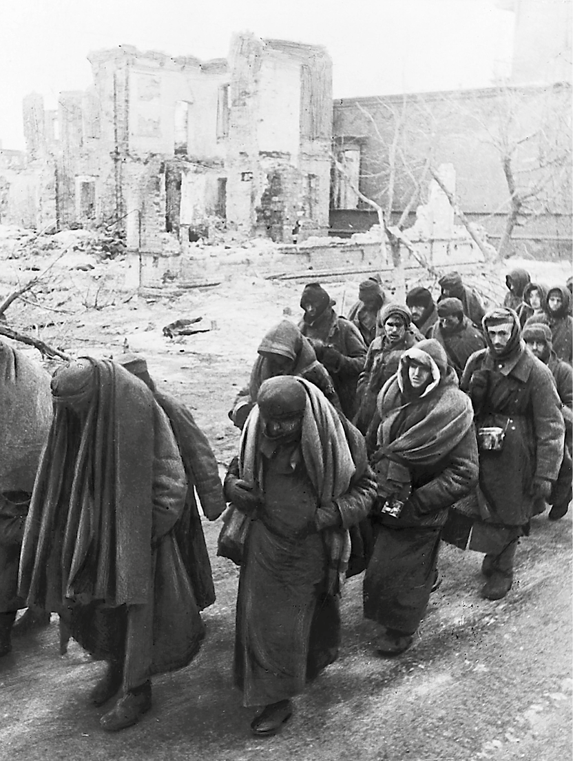 Fotografia em preto e branco. Homens andando em fila. Estão agasalhados com grandes casacos e mantas sobre as costas. Ao fundo, ruínas de casas.