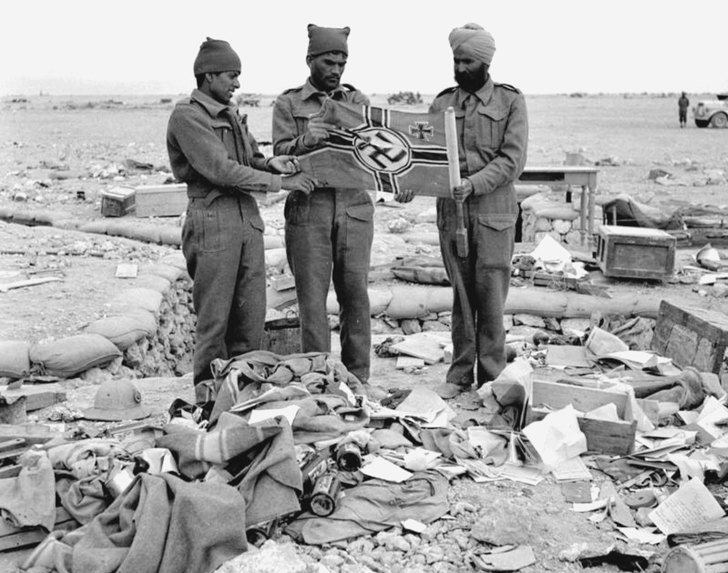 Fotografia em preto e branco. Três soldados em meio a escombros. Dois usam gorro e um usa um turbante na cabeça. Seguram nas mãos uma bandeira nazista.