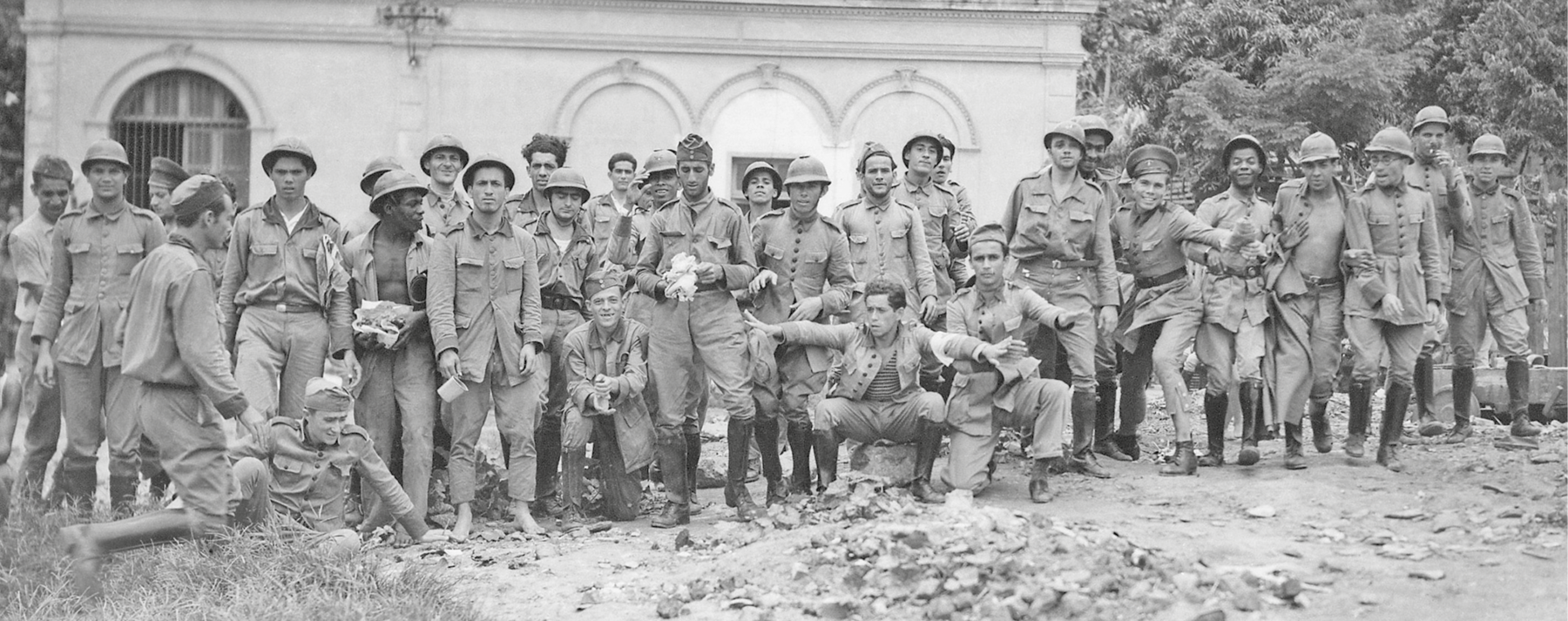 Fotografia em preto e branco. Grupo de homens posando para foto. Estão descontraídos. Todos vestem roupas militares, a maioria usa capacete.