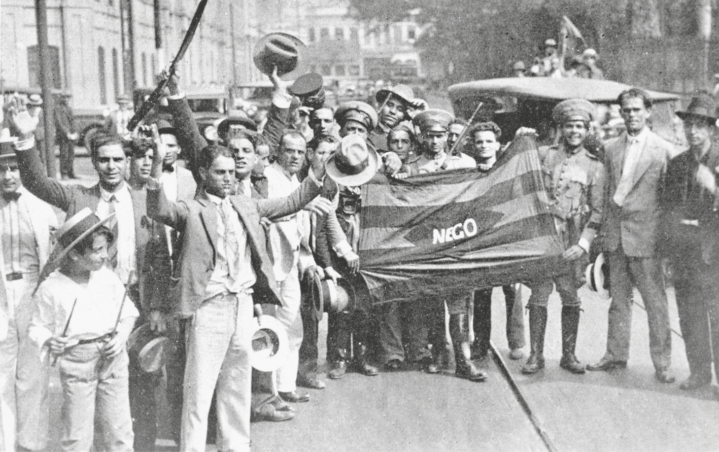 Fotografia em preto e branco. Manifestantes na rua, agrupados, segurando uma bandeira com listras horizontais e a palavra NEGO no centro. Alguns estão com os braços erguidos.
