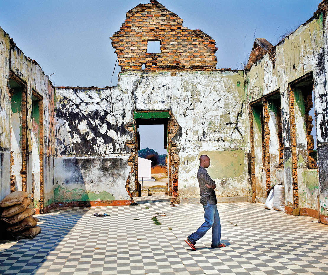 Fotografia. Homem dentro de uma construção em ruínas. O piso é xadrez e as paredes esverdeadas.