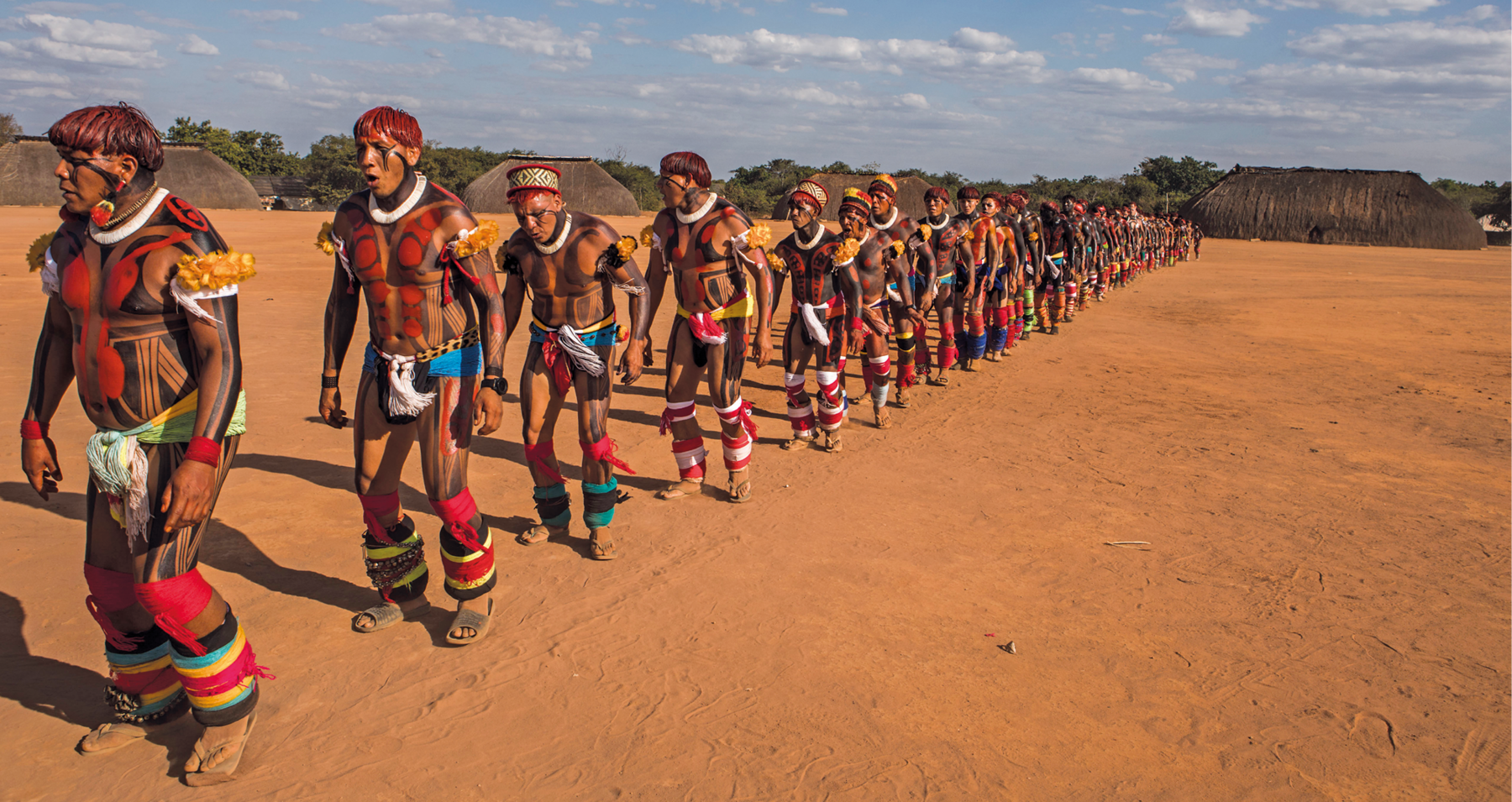 Fotografia. Indígenas alinhados em uma longa fila sobre chão de terra. Estão com o cabelo pintado de vermelho e desenhos ornamentando o corpo. Usam adereços nos braços e nas pernas. Ao fundo, várias ocas.