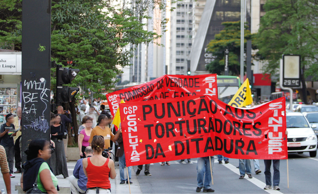 Fotografia. Grupo de pessoas em uma manifestação em avenida. Carregam uma grande faixa vermelha com os dizeres: PUNIÇÃO AOS TORTURADORES DA DITADURA.
