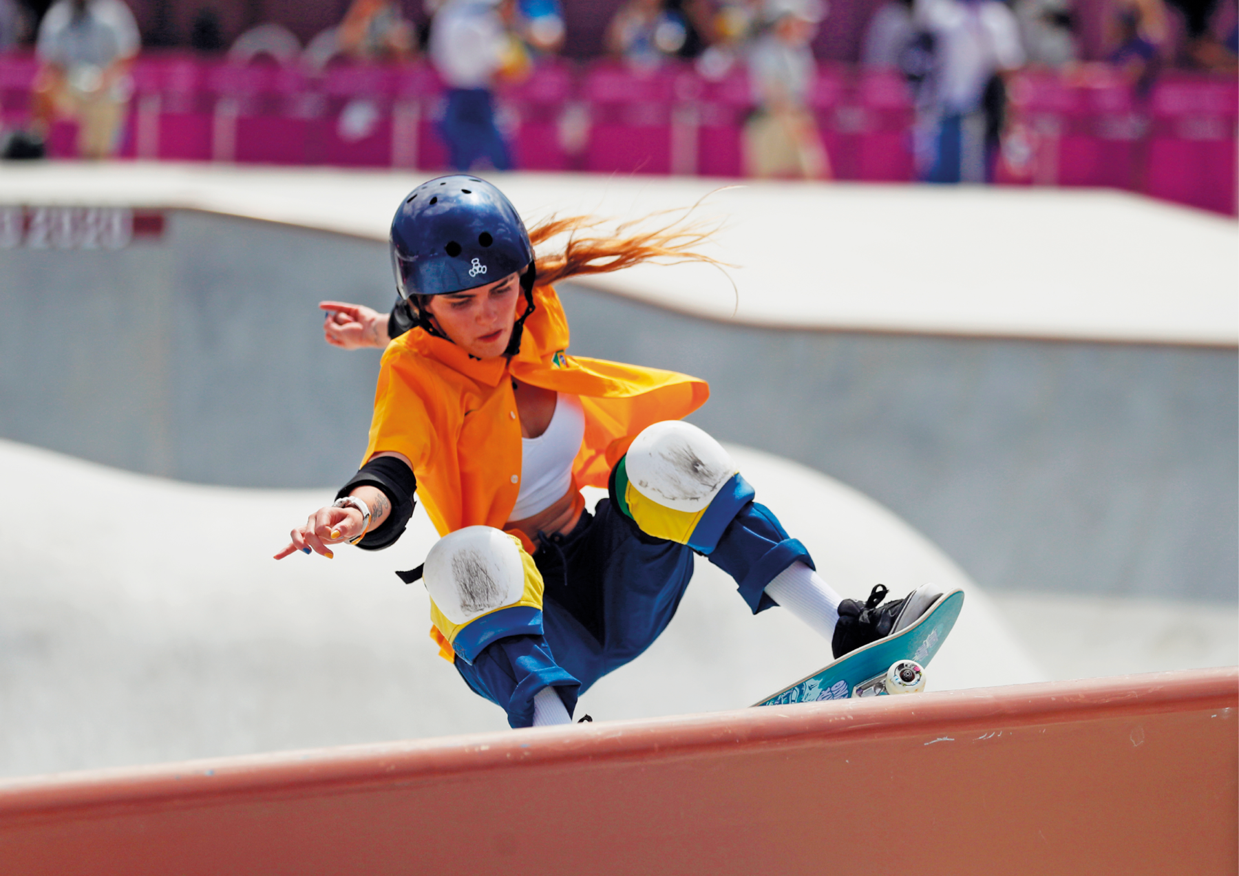 Fotografia. Menina skatista de capacete e joelheiras fazendo uma manobra. Está com as pernas flexionadas e um dos braços para trás. Usa camisa laranja e calça azul.