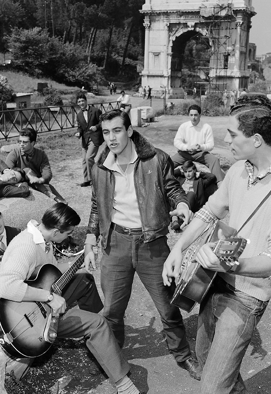 Fotografia em preto e branco. Jovens de camisa, calça jeans e cabelos penteados com topete. Tocam instrumentos ao ar livre. Ao fundo, ruínas de uma construção do Império Romano.