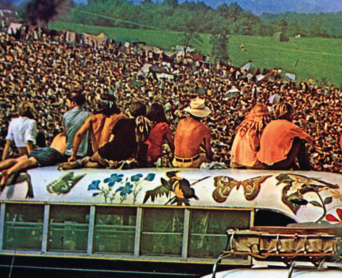 Fotografia. Grupo de pessoas sentado no teto de um ônibus. Ao fundo, uma multidão em uma colina. O ônibus tem o teto branco com desenhos de flores e animais.