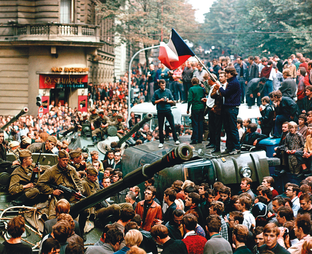 Fotografia. Manifestação de estudantes aglomerados em uma rua em meio a tanques de guerra. Alguns deles estão em pé, sobre um veículo. Um deles segura uma bandeira da Tchecoslováquia. No canto esquerdo da imagem e ao fundo, militares observam a cena.
