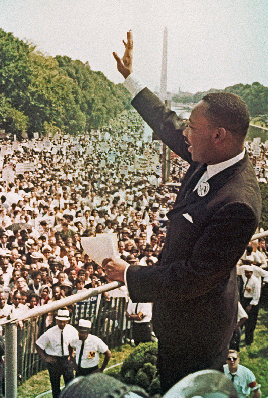 Fotografia. Martin Luther King Junior, homem negro, vestindo terno e gravata. Está no topo de uma colina acenando para uma multidão. Segura um papel com uma das mãos.