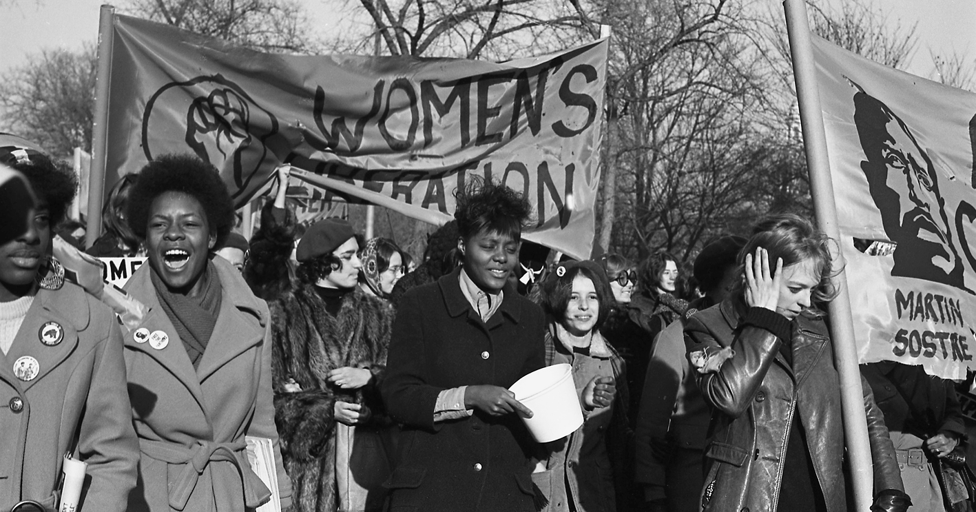 Fotografia em preto e branco. Mulheres negras e brancas reunidas em uma manifestação na rua. Carregam faixas.