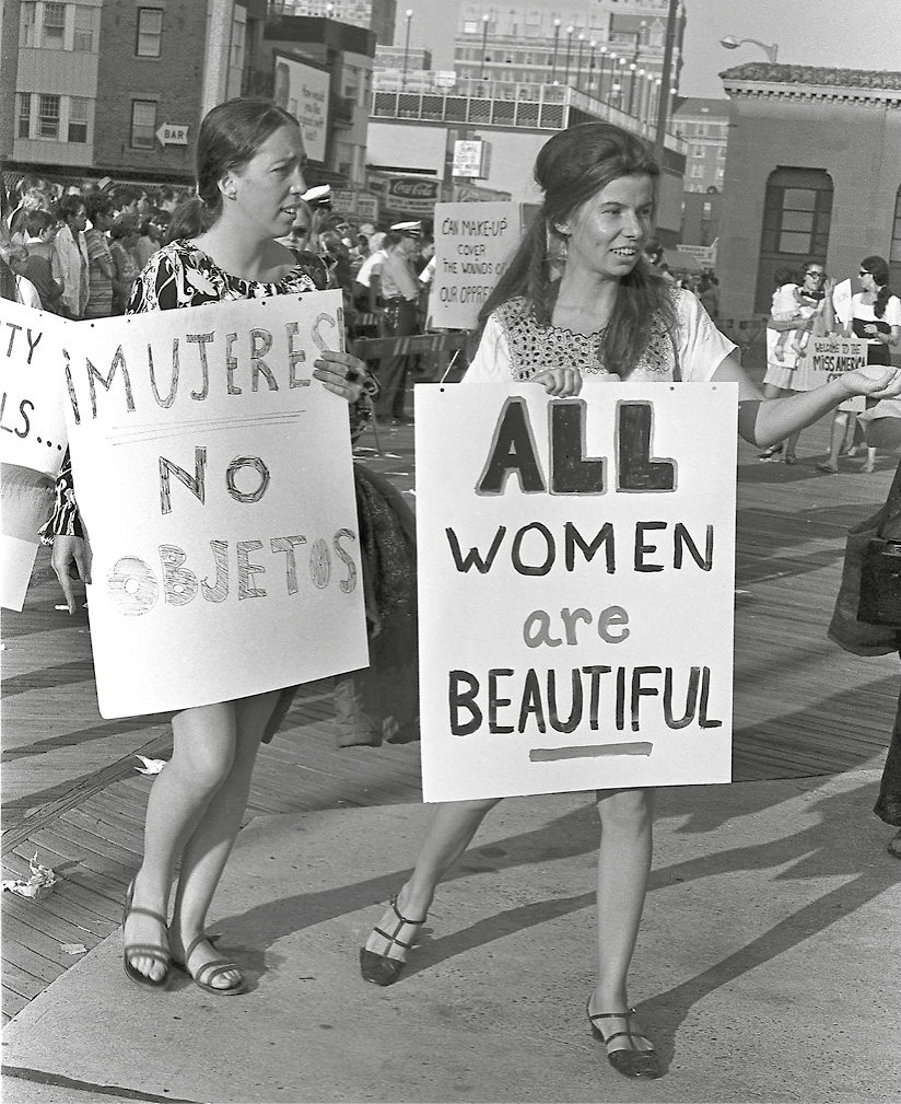 Fotografia em preto e branco. À frente, duas mulheres segurando cartazes com palavras de ordem. Ao fundo, outras pessoas na manifestação.