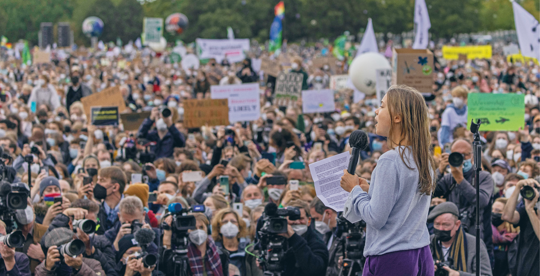 Fotografia. Greta Thunberg, moça loira de cabelo comprido discursando em um palanque. À frente dela, fotógrafos, repórteres e uma multidão com cartazes.