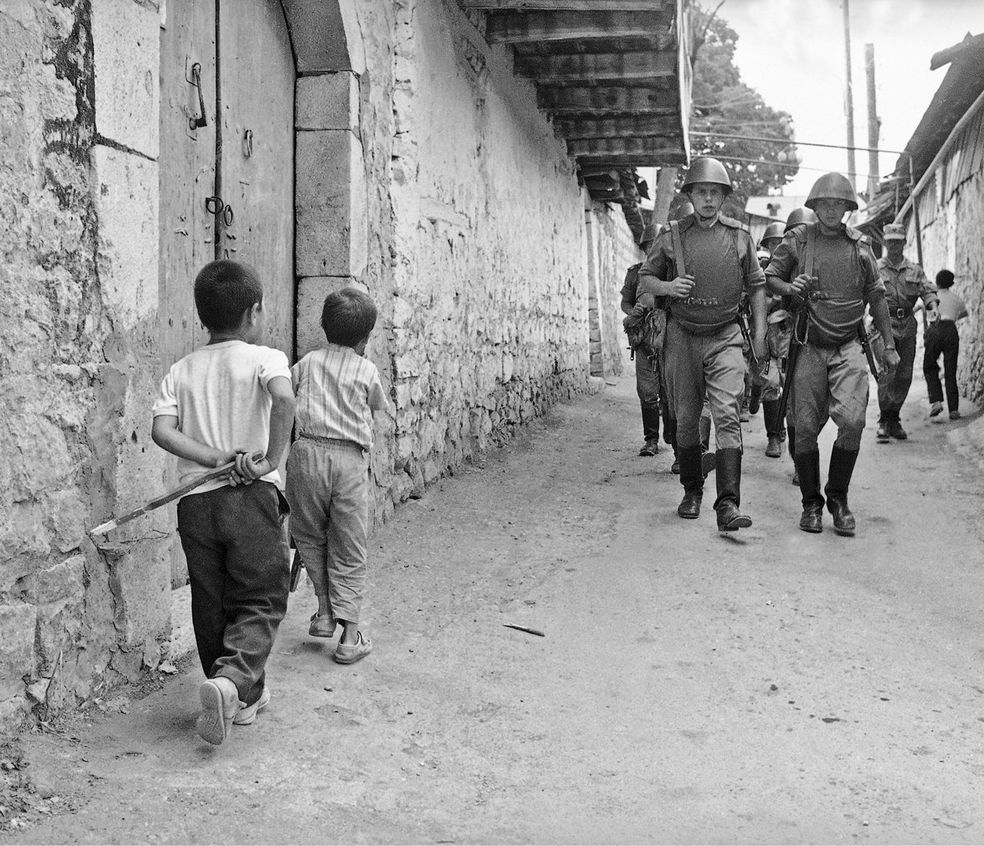 Fotografia em preto e branco. Militares passando por crianças na rua de um vilarejo.