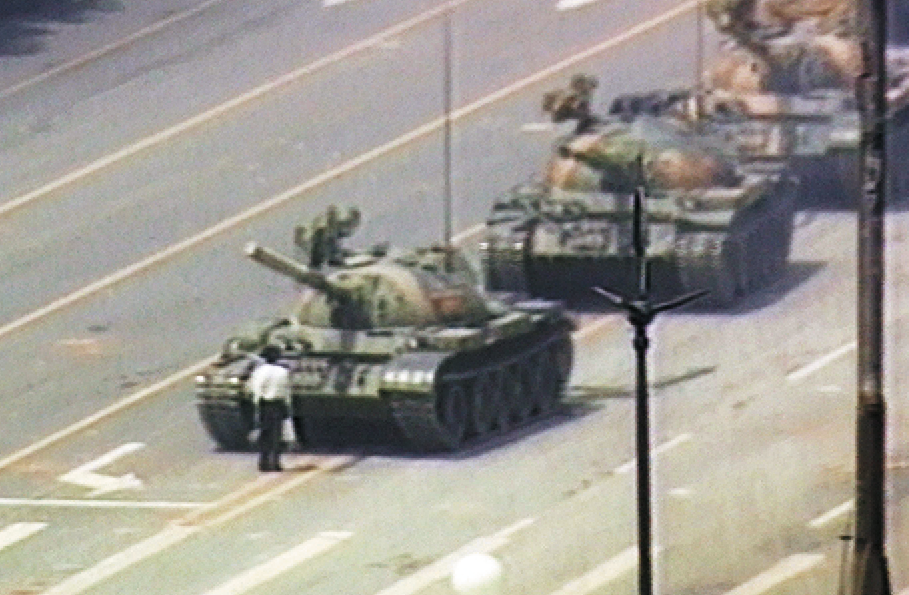 Fotografia. Em uma grande avenida, pessoa em pé, impedindo o fluxo de tanques de guerra que estão à sua frente.
