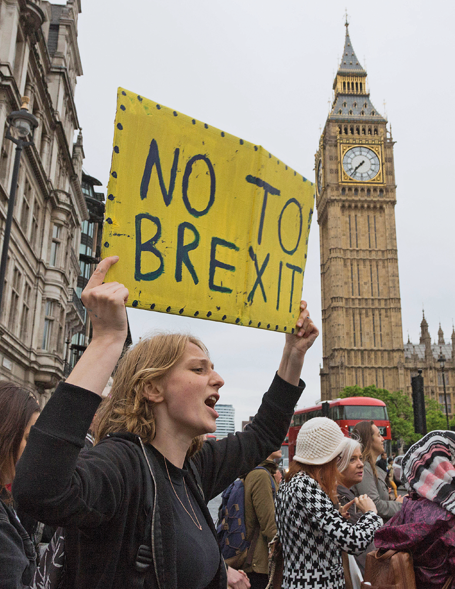 Fotografia. Jovens em uma manifestação. À frente um cartaz feito à mão com os dizeres cuja tradução é: Não ao Brexit. Ao fundo, uma grande torre com um relógio.