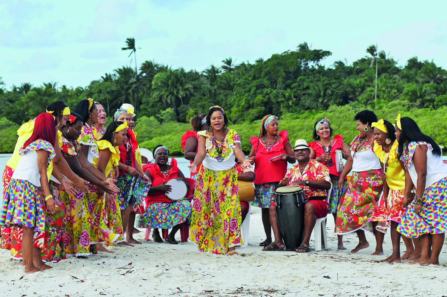 Fotografia. Mulheres na praia, em uma roda de samba. Vestem saias longas e coloridas. Usam faixas e adornos nos cabelos. Algumas estão sentadas tocando instrumentos ao lado de um homem de chapéu. Ao redor, outras mulheres dançam.