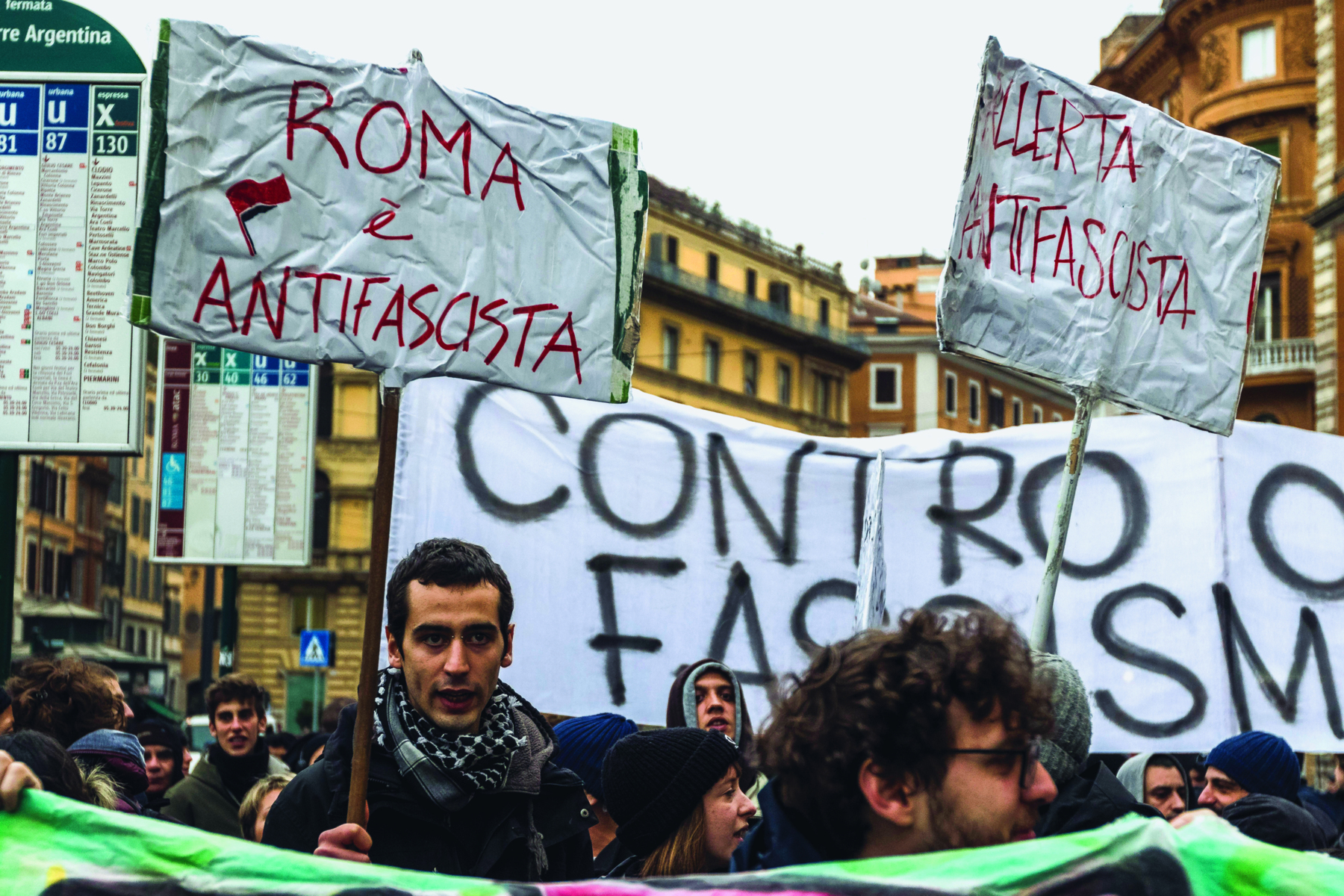 Fotografia. Pessoas na rua em um protesto. Carregam cartazes e faixas escritos à mão. À frente um rapaz carrega um cartaz com os dizeres: ROMA É ANTIFASCISTA.