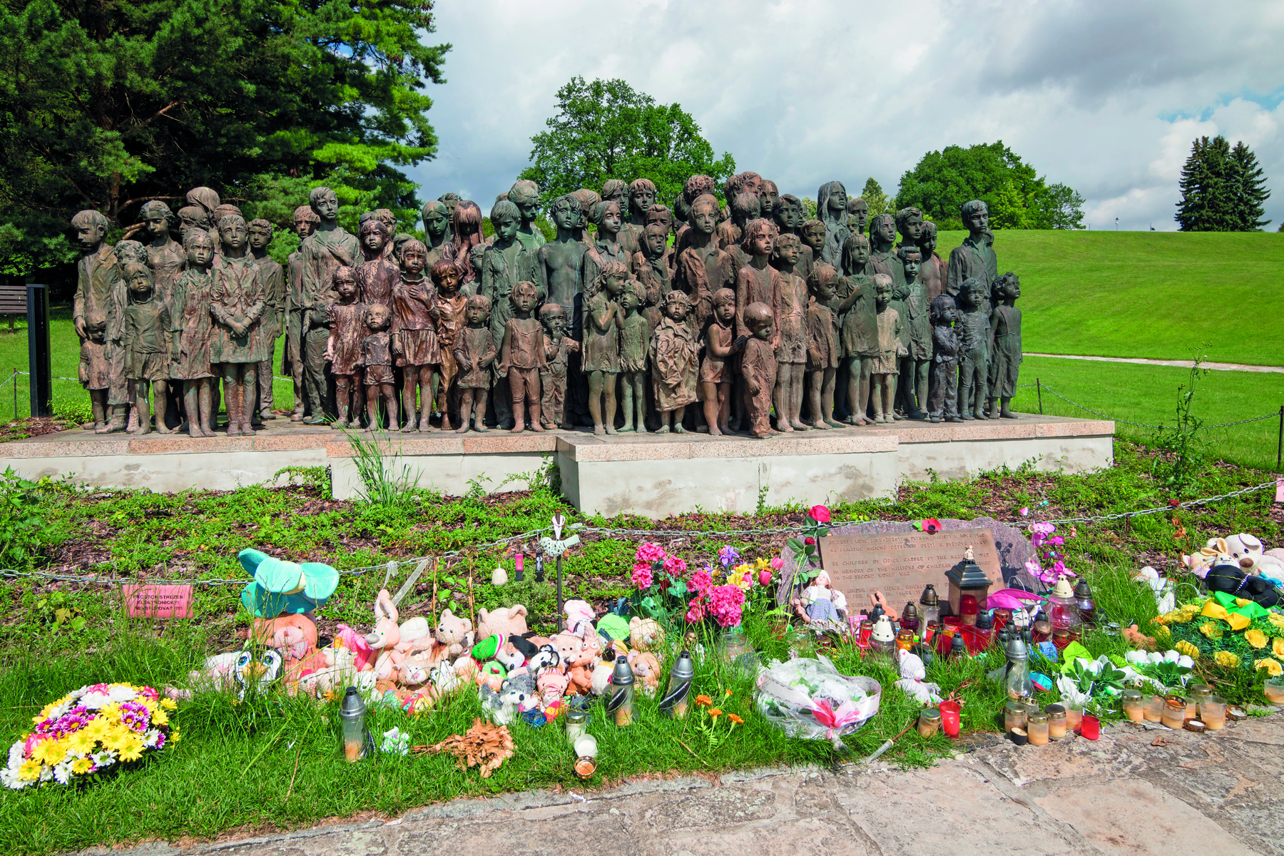Fotografia. Monumento de bronze em um campo gramado, composto por diversas estátuas de crianças, de idades variadas. À frente do monumento, flores, pelúcias e velas.