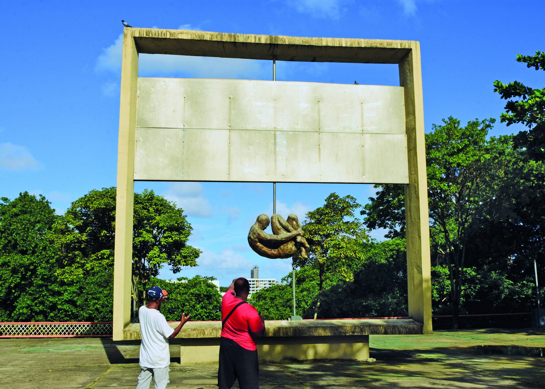 Fotografia. Dois homens observam uma escultura em uma região arborizada. Moldura retangular, com uma placa retangular disposta horizontalmente no centro. Abaixo da placa, uma pessoa com o corpo encolhido, abraçando as pernas.