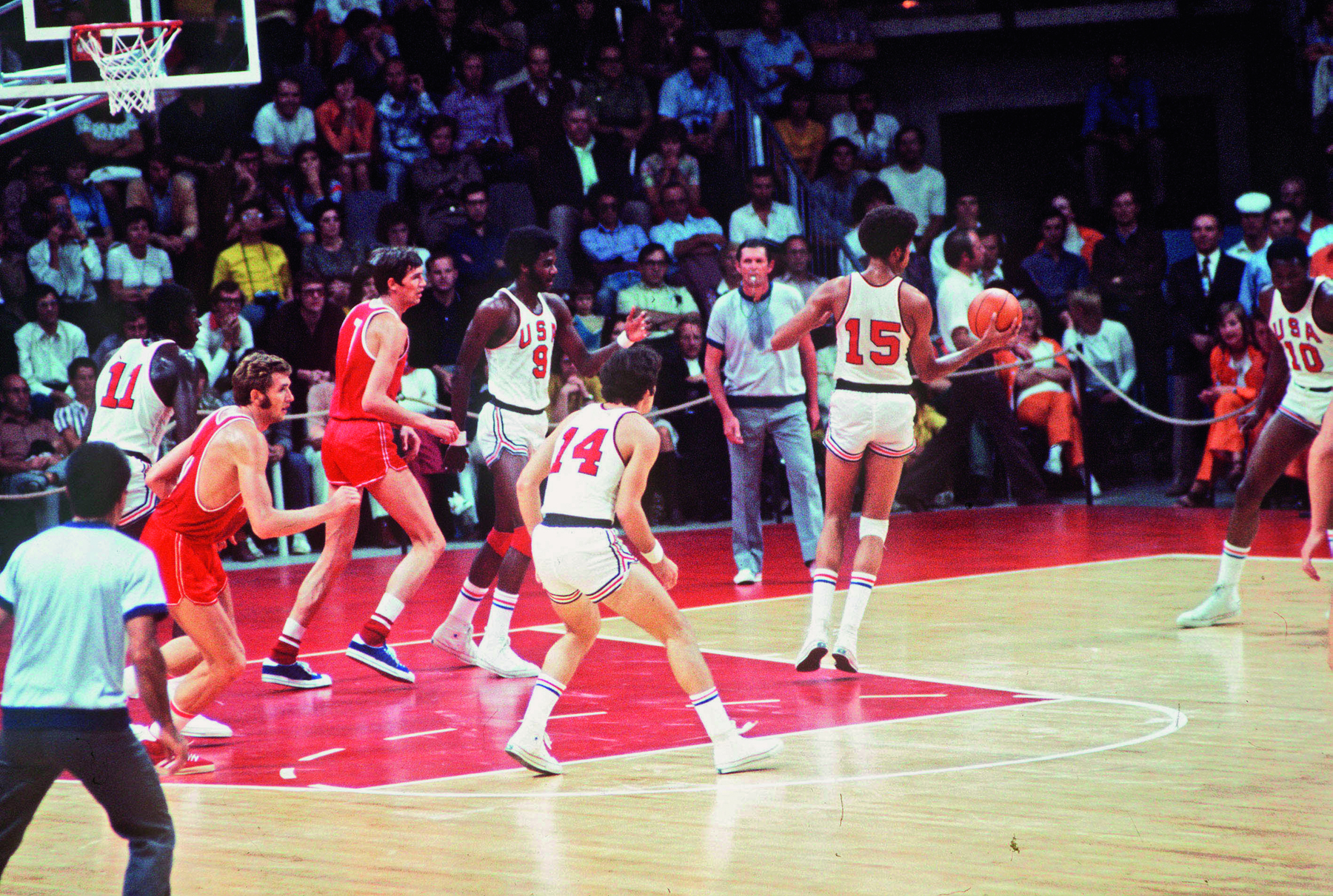 Fotografia. Pessoas assistem a uma partida de basquete. O time dos Estados unidos usa uniforme branco com números vermelhos. O time da união Soviética usa uniforme vermelho com números em brancos. Os jogadores tem as pernas e braços compridos.