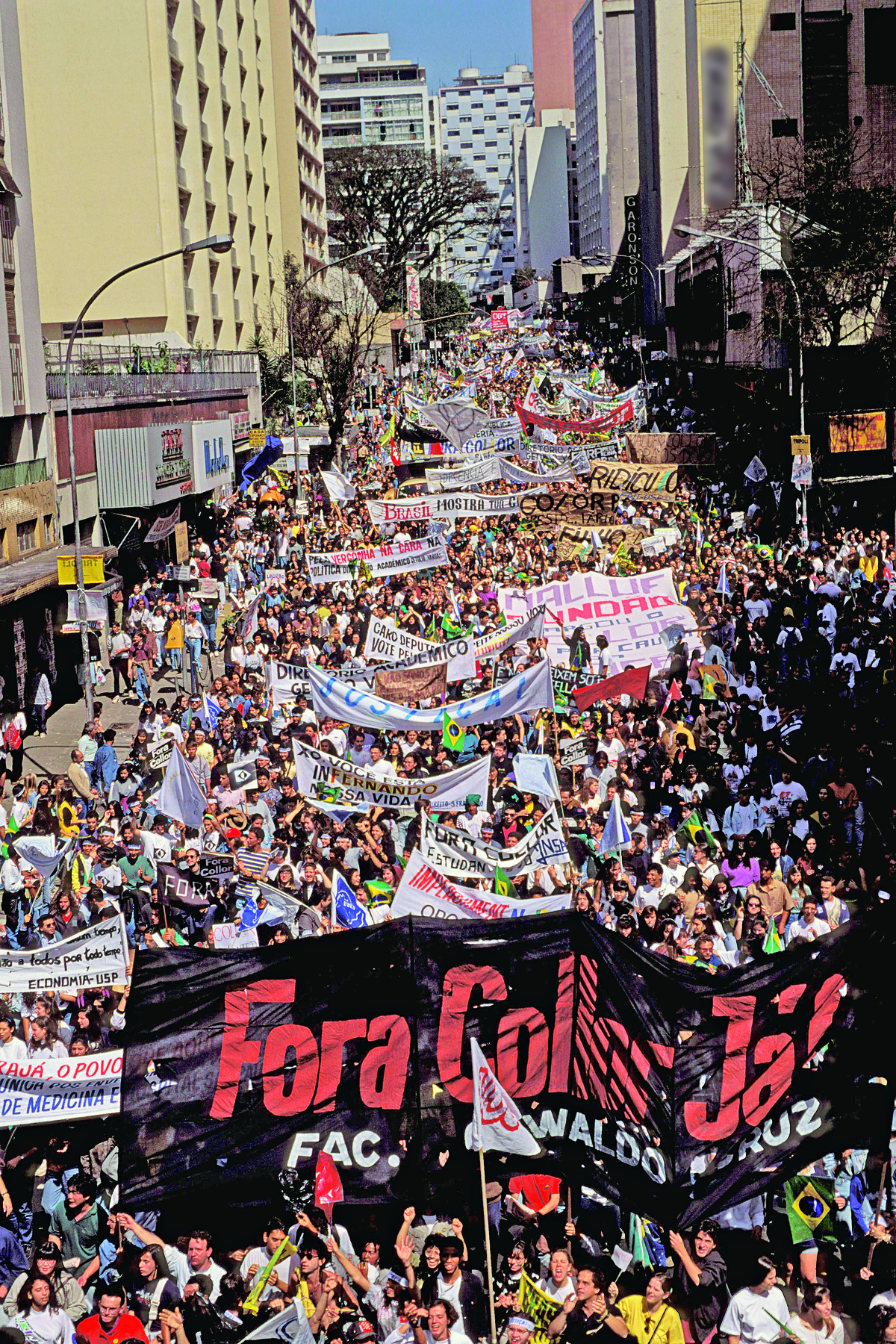 Fotografia. Multidão na rua em uma manifestação. Carregam muitos cartazes e faixas. À frente, a faixa tem os dizeres: FORA COLLOR JÁ!