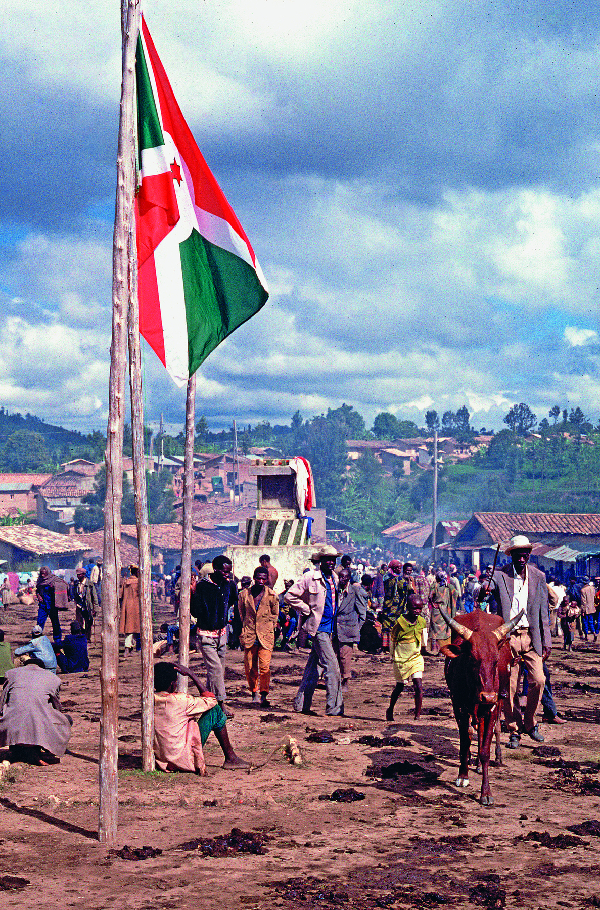 Fotografia. Pessoas caminhando sobre chão de terra. À frente, uma bandeira no mastro. Ao fundo, um vilarejo.