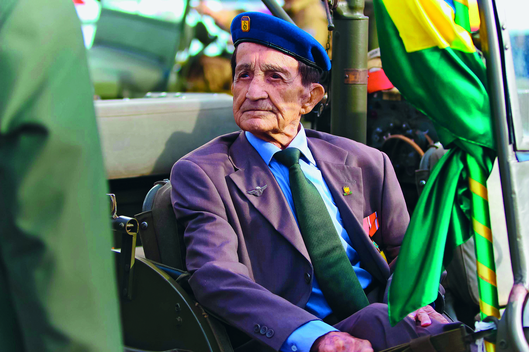 Fotografia. Nestor da Silva, senhor de terno e boina azul com o símbolo da FEB na frente. Está sentado perto de um mastro com uma bandeira do Brasil enrolada.