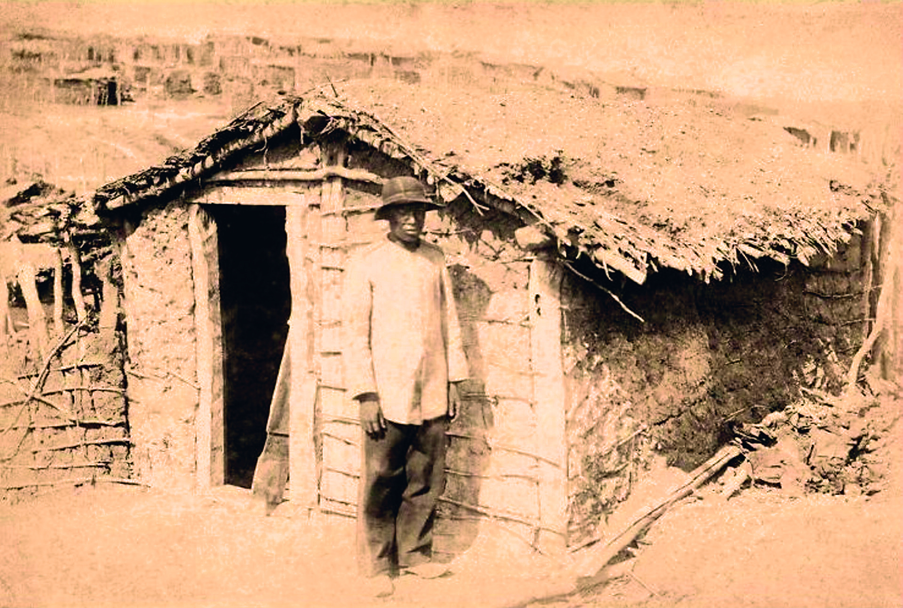 Fotografia em sépia. Homem em pé, vestindo calça e camisa de manga longa. Usa um chapéu arredondado. Está em frente a uma casa rústica com uma pequena porta e telhado de palha.