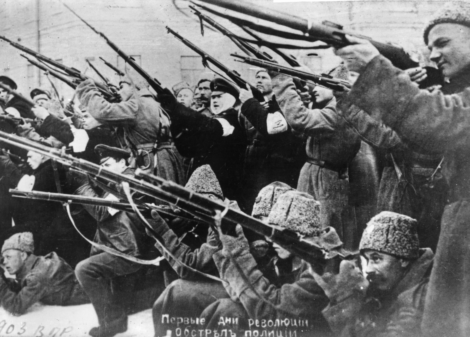 Fotografia em preto e branco. Homens de sobretudo agrupados empunhando  armas de fogo de cano longo para a mesma direção.