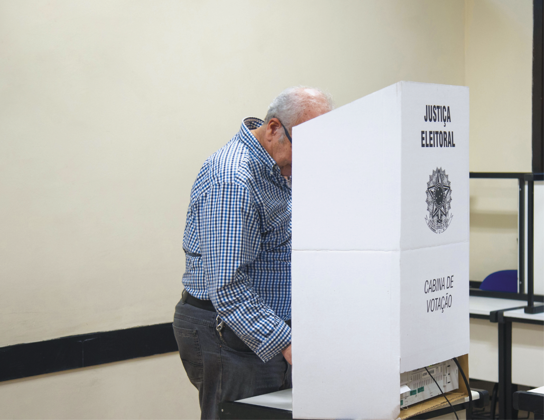 Fotografia. Senhor em pé, votando. Ele usa camisa xadrez azul, preta e branca, e calça escura. Está atrás da Cabine de Votação, biombo de papelão branco com o emblema da Justiça Eleitoral.