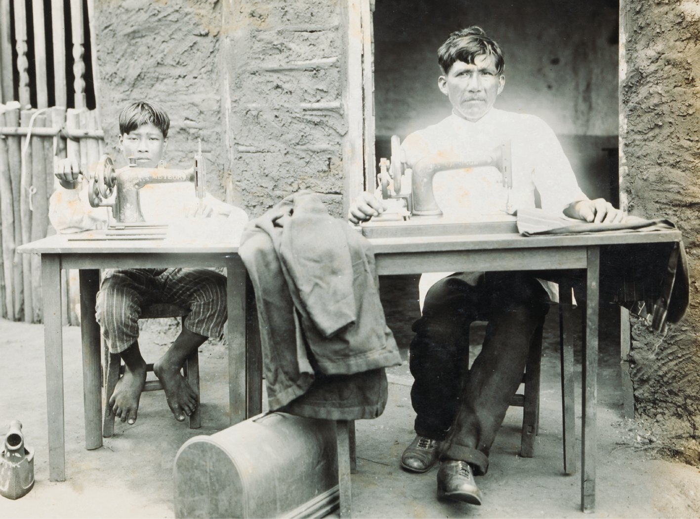 Fotografia em preto e branco. Dois indígenas, um homem e um menino, sentados trabalhando, cada um em uma máquina de costura. O menino usa camisa clara e calça listrada, e está descalço. O homem usa camisa clara, calça social escura e sapato preto.
