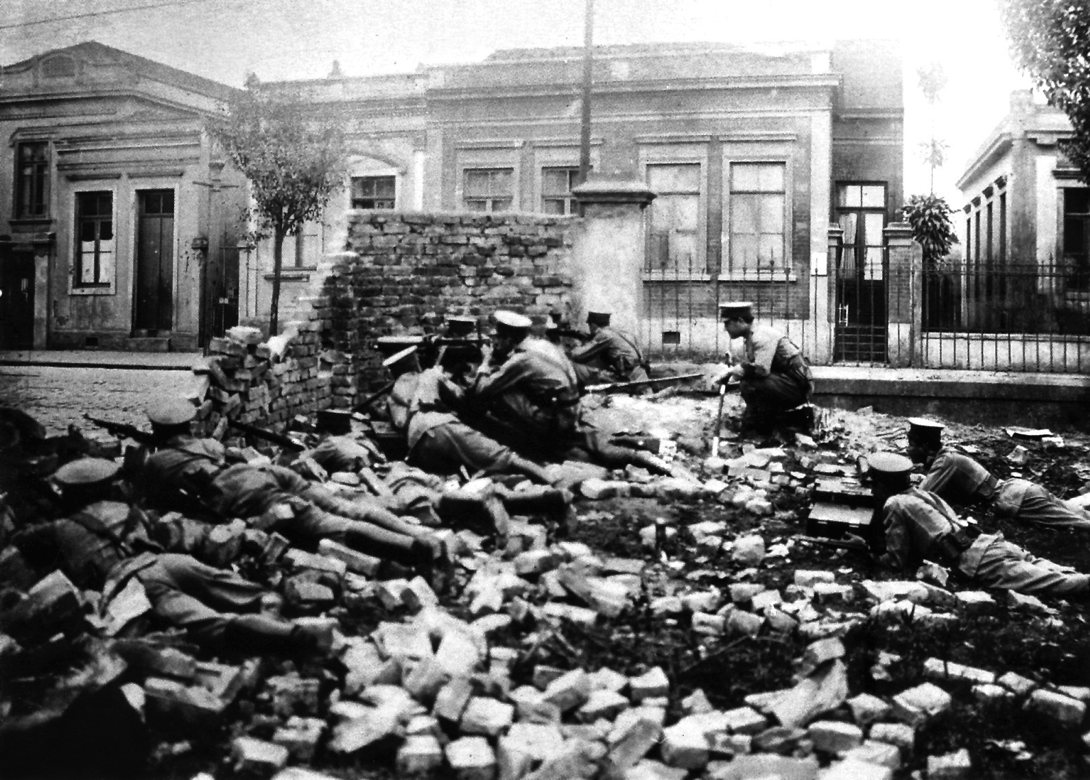 Fotografia em preto e branco. Soldados abaixados em uma trincheira feita de escombros de um muro. Alguns estão deitados com as armas apontadas. Ao fundo algumas casas.