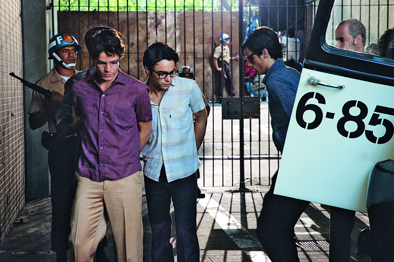 Cena de filme. Três rapazes de óculos, calça e camisa social, presos pela polícia, descem de um camburão. Policiais observam. Ao fundo, um grande portão fechado.