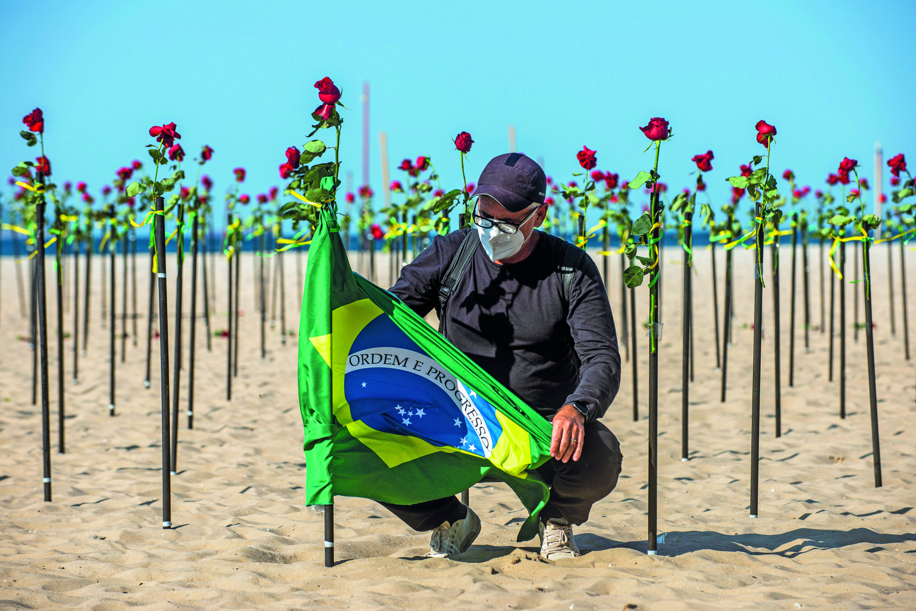 Fotografia. Um homem prende a bandeira do Brasil em uma estaca fincada na areia da praia. Ele usa boné e roupas escuras, e máscara de proteção facial. Atrás dele, dezenas de rosas em estacas encaixadas na areia.