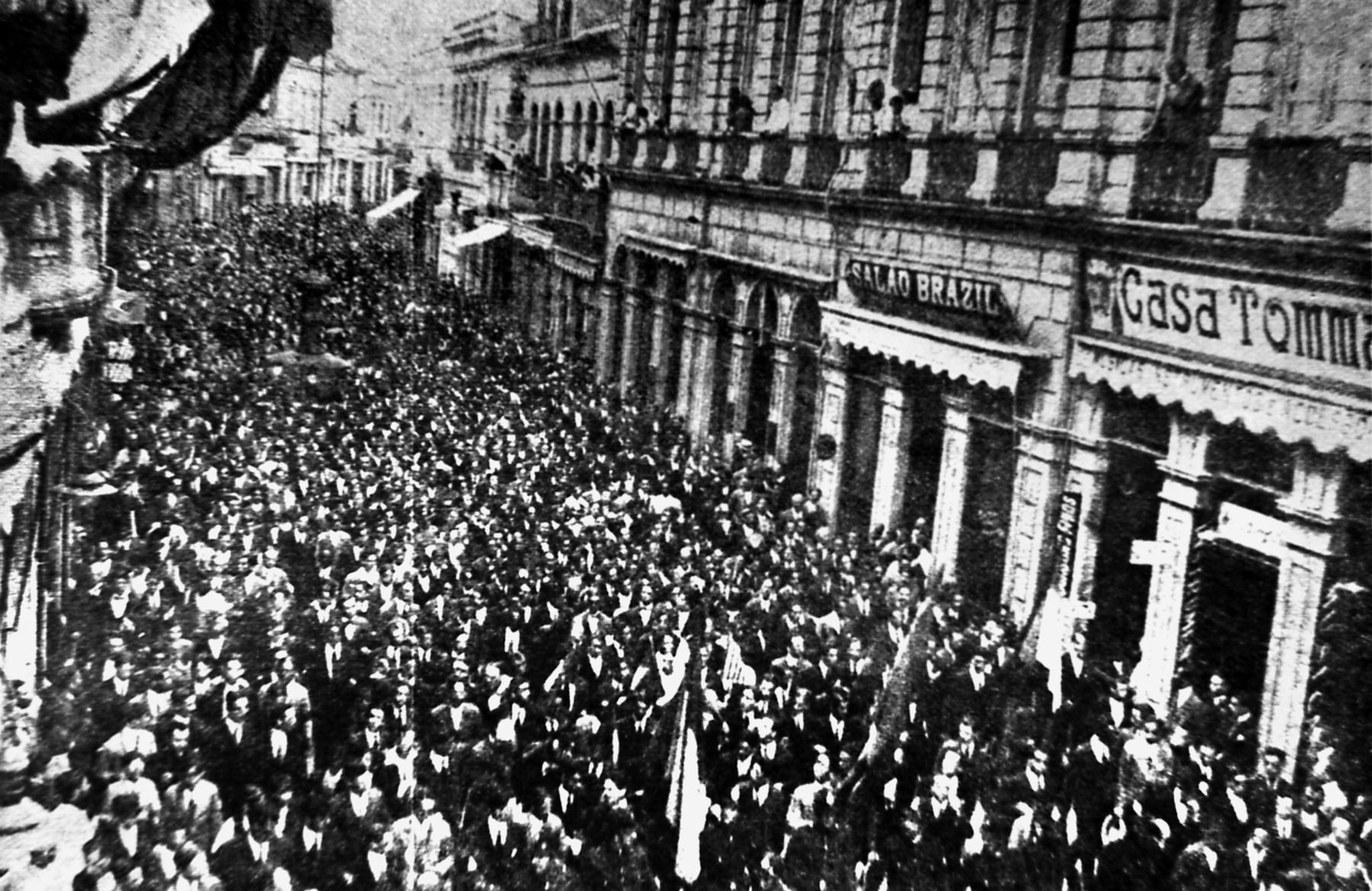 Fotografia em preto e branco. Multidão aglomerada na rua em um protesto. Nas laterais as entradas de prédios.