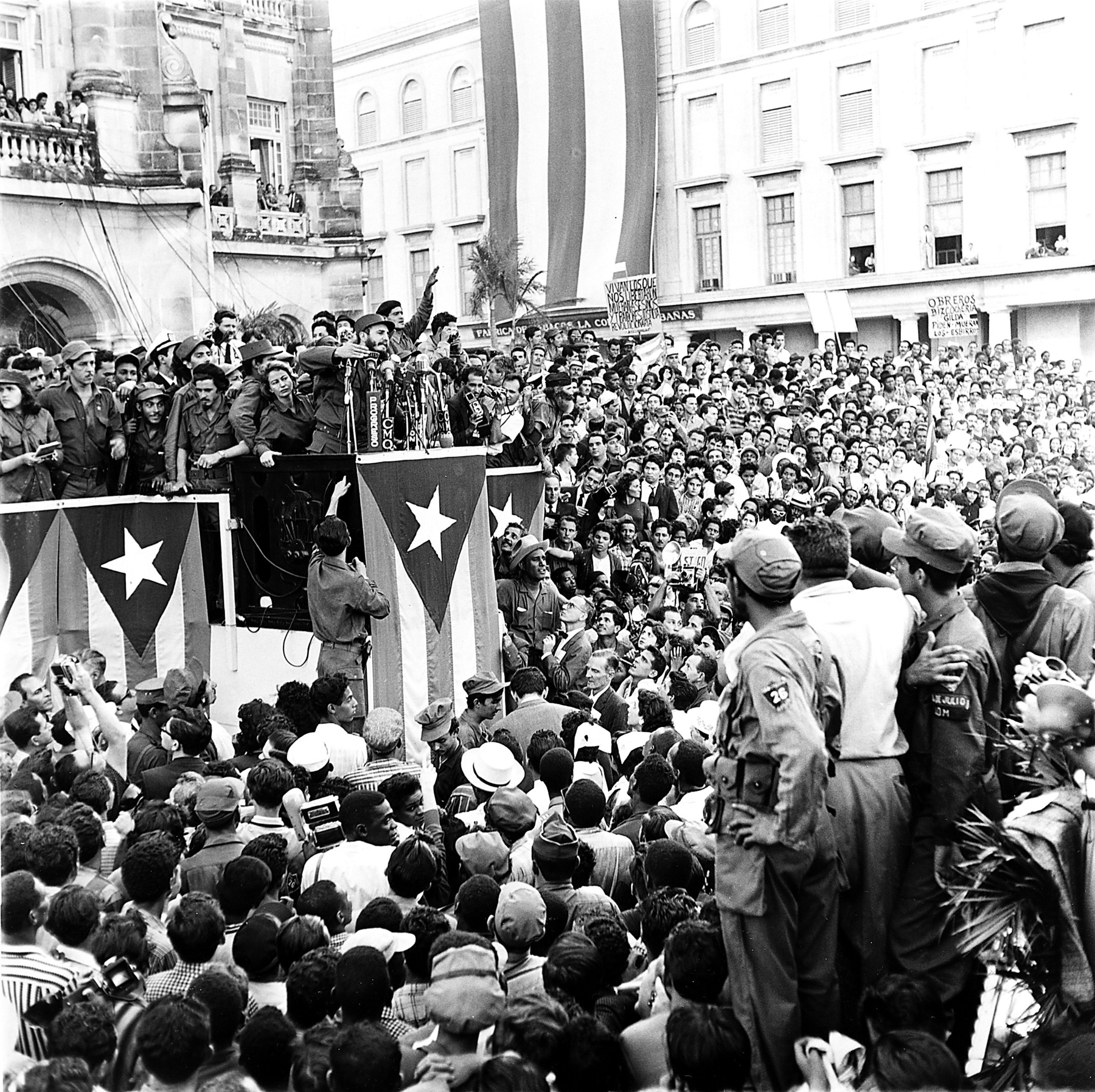 Fotografia em preto e branco. Fidel Castro, homem de barba cheia e boina. Está sobre um palanque ao lado de diversas pessoas. Ao redor uma multidão o observa. Na parte de baixo do palanque, várias bandeiras de Cuba dependuradas.