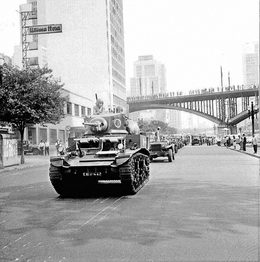 Fotografia em preto e branco. Tanques de guerra se deslocando em uma rua. Ao fundo, prédios e um viaduto.