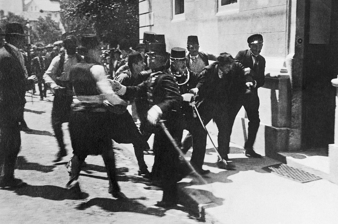 Fotografia em preto e branco. Diversos militares aglomerados  ao redor de Gavrilo Princip, rapaz de casaco e boina pretos. Estão na porta de um edifício.