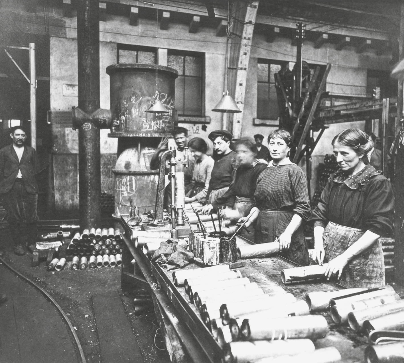 Fotografia em preto e branco. Mulheres em uma linha de produção. Estão em pé atrás de uma longa bancada baixa. Sobre ela diversos tubos. Ao fundo, grandes dutos interligados. Atrás e à esquerda, alguns homens observam.