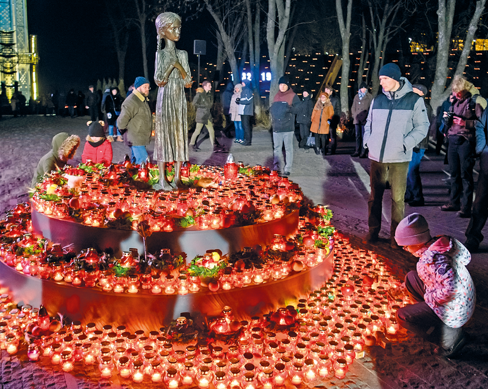 Fotografia. Pessoas agasalhadas com casacos e gorros estão perto da estátua de uma menina. Ao redor dela diversas velas vermelhas acesas.