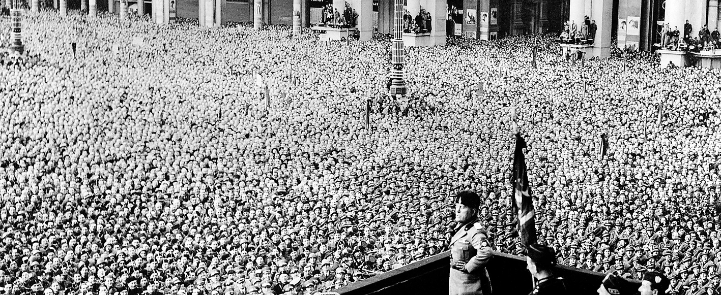 Fotografia em preto e branco. Em primeiro plano, Benito Mussolini com seu traje militar, em pé com as mãos na cintura em um palanque. Ao redor, multidão aglomerada.