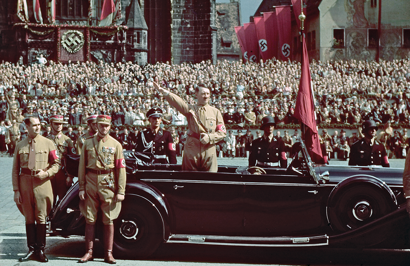 Fotografia. Adolf Hitler, homem de cabelos escuros penteados de lado e bigode escuro e curto. Uma das mãos está apoiada no cinto e a outra está esticada diagonalmente para frente, em uma saudação. Ele veste um macacão bege com uma braçadeira vermelha com a suástica desenhada. Está em pé, dentro de um carro. Perto dele, alguns militares. Ao fundo, uma multidão aglomerada e diversas bandeiras com a suástica  nazista.