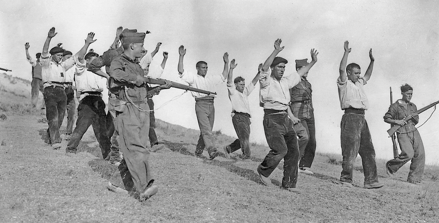 Fotografia em preto e branco. Militares apontando armas para um grupo de homens em uma colina. Eles estão com os braços erguidos.