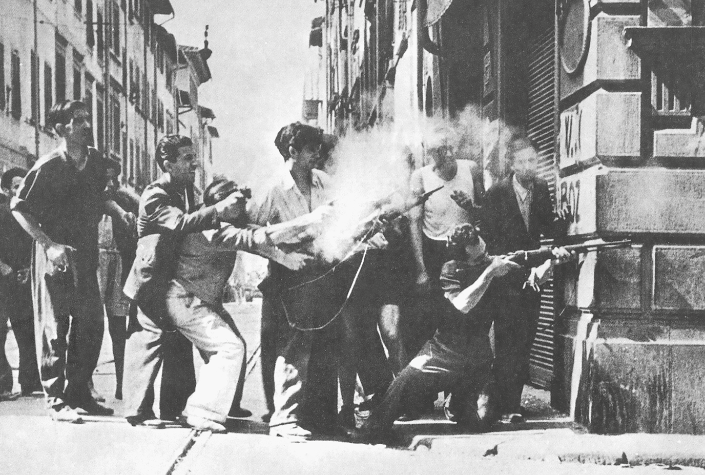 Fotografia em preto e branco. Homens empunhando armas em uma esquina. Um deles está ajoelhado, apontando a arma, com o corpo parcialmente atrás da parede de uma construção. Outros observam.