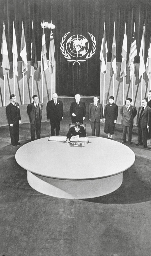 Fotografia em preto e branco. Homens vestidos socialmente, em pé, ao redor de uma grande mesa oval. Um homem está sentado à mesa. Sobre ela, alguns papéis. Ao fundo, diversas bandeiras em mastros e o símbolo da ONU na parede.